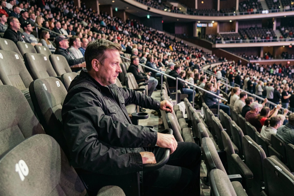 Homme assis dans les gradins numérotés d'une salle de concert type Bercy, vue sur la fosse et la scène lors d'un concert rock